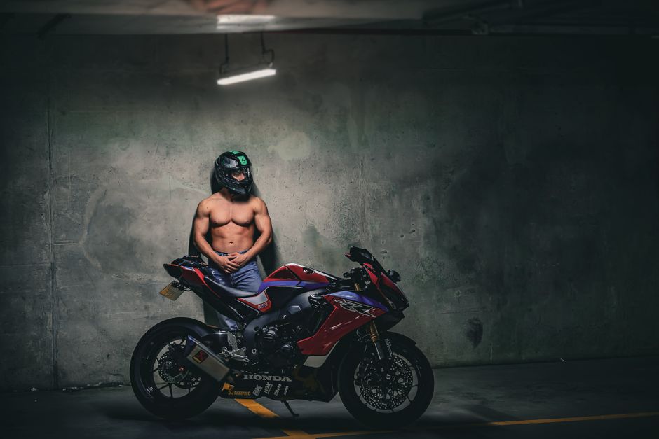 A motorcyclist in a helmet stands beside a sports bike in a dimly lit urban garage.