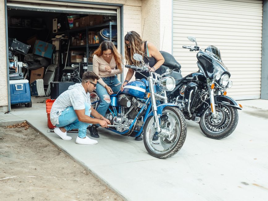 Three individuals repairing motorcycles outside a garage on a sunny day.