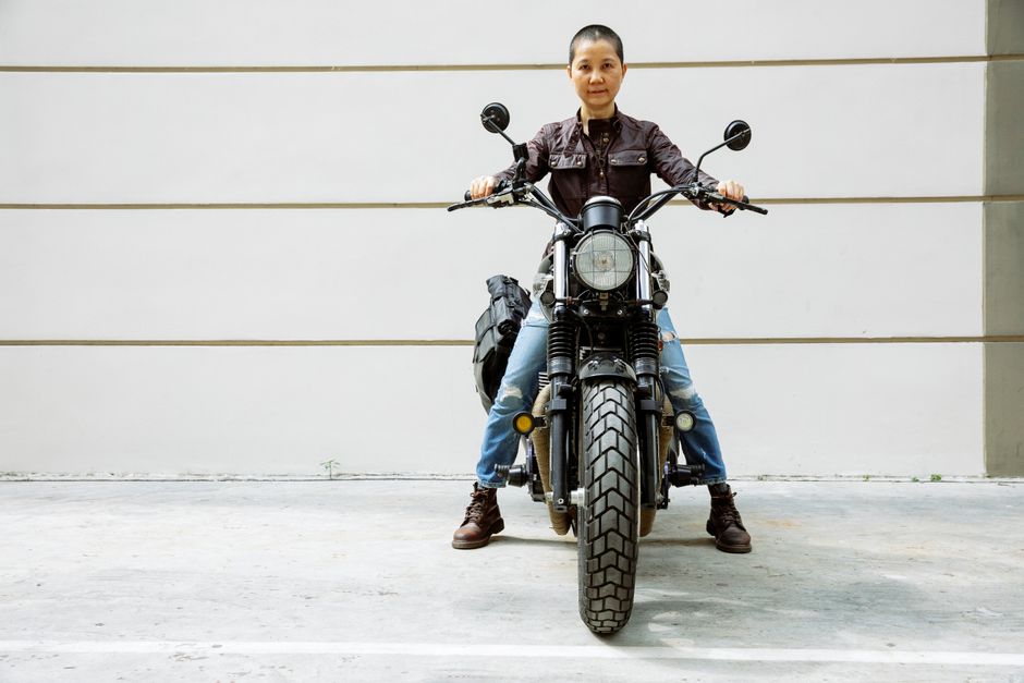 Serious mature Asian female biker in jacket and jeans sitting on black motorcycle on asphalt road in street near wall and looking at camera in daytime