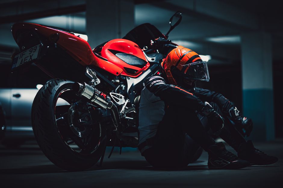A motorcyclist in full gear relaxes beside a red motorcycle in a dimly lit garage.