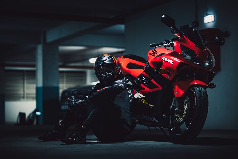 Motorcyclist in full gear sits in a dimly lit garage next to a vibrant red sport bike, creating a dramatic atmosphere.
